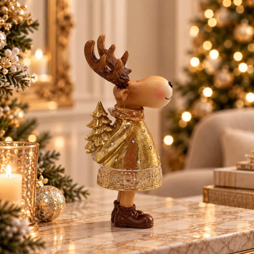 A festive Christmas reindeer figurine wearing a glittery gold dress and brown boots, standing on a marble surface in a warmly lit holiday room with blurred Christmas trees and candles in the background.