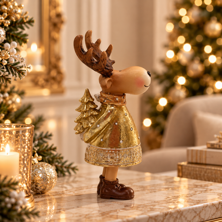 A festive Christmas reindeer figurine wearing a glittery gold dress and brown boots, standing on a marble surface in a warmly lit holiday room with blurred Christmas trees and candles in the background.